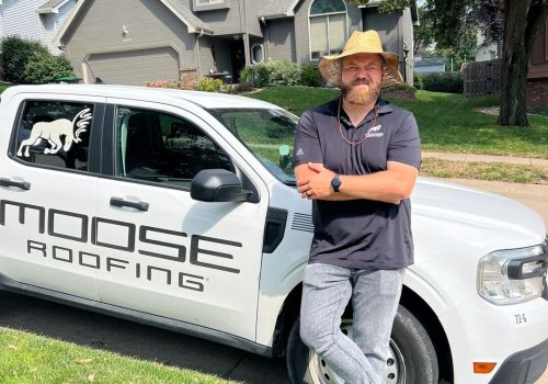 Man in straw hat stands by Moose Roofing truck in suburban neighborhood.