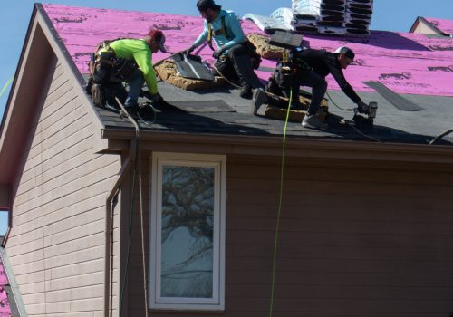 Roofers work on a house, installing shingles under a clear blue sky.