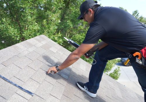 Roofer inspects shingles on a residential roof surrounded by trees.