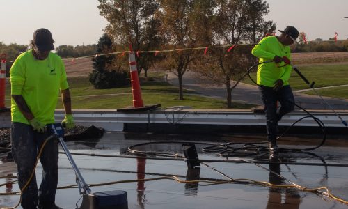 Roofers in bright shirts work on a flat commercial roof under clear skies.