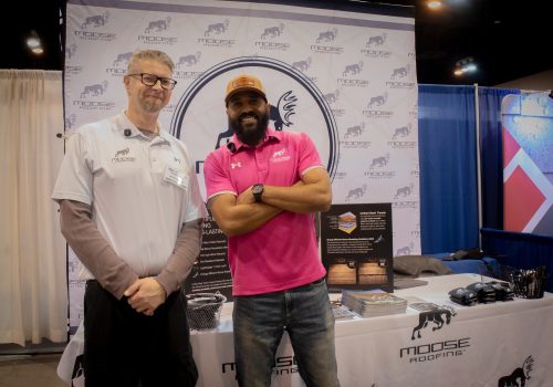 Two men stand at a roofing business booth with branded materials displayed.