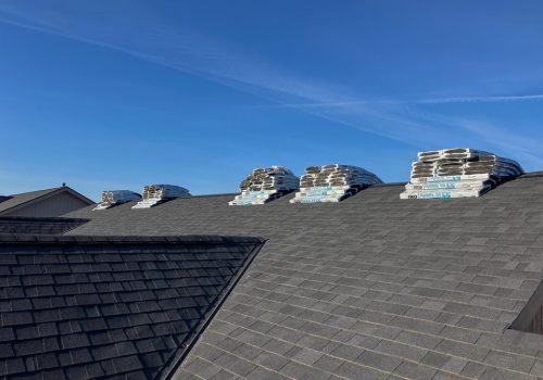 Stacks of shingles are arranged on top of a grey shingle roof under a clear sky.