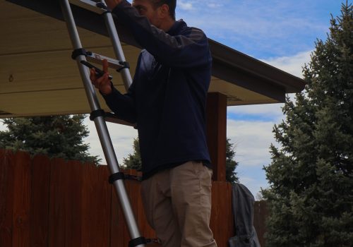 Man on ladder inspecting roof with clear blue sky background and green trees.