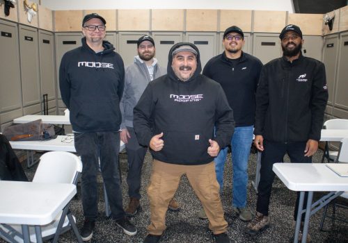 Roofing team posing in a training room with tables and chairs visible.