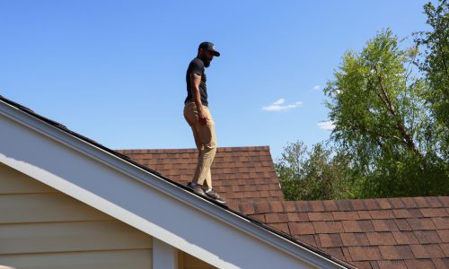 A professional inspects a residential roof under a clear blue sky.