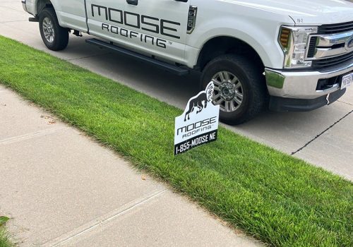 White truck with "Moose Roofing" logo parked on grass. Nearby sign displays company name and phone number.